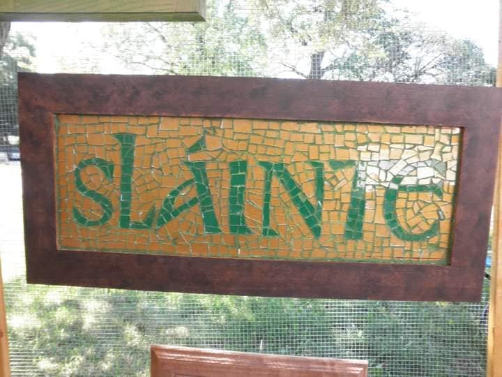 Mockzaic of Irish cheer Sláinte, in green with yellow background and brown rustic wooden frame hanging in window with trees in the background.
